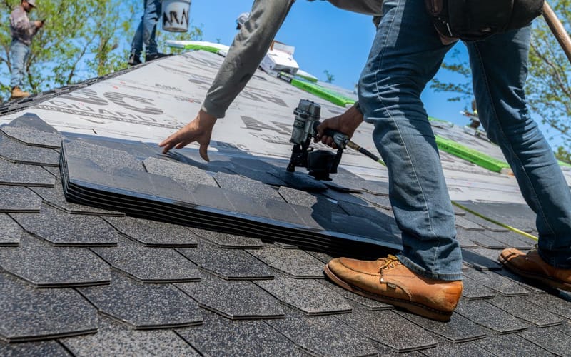 Roofer fastening new asphalt architectural shingles with a nail gun during a roof replacement