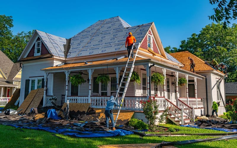 Roof replacement in progress with old shingles torn off, exposed decking, and debris collected on tarps below